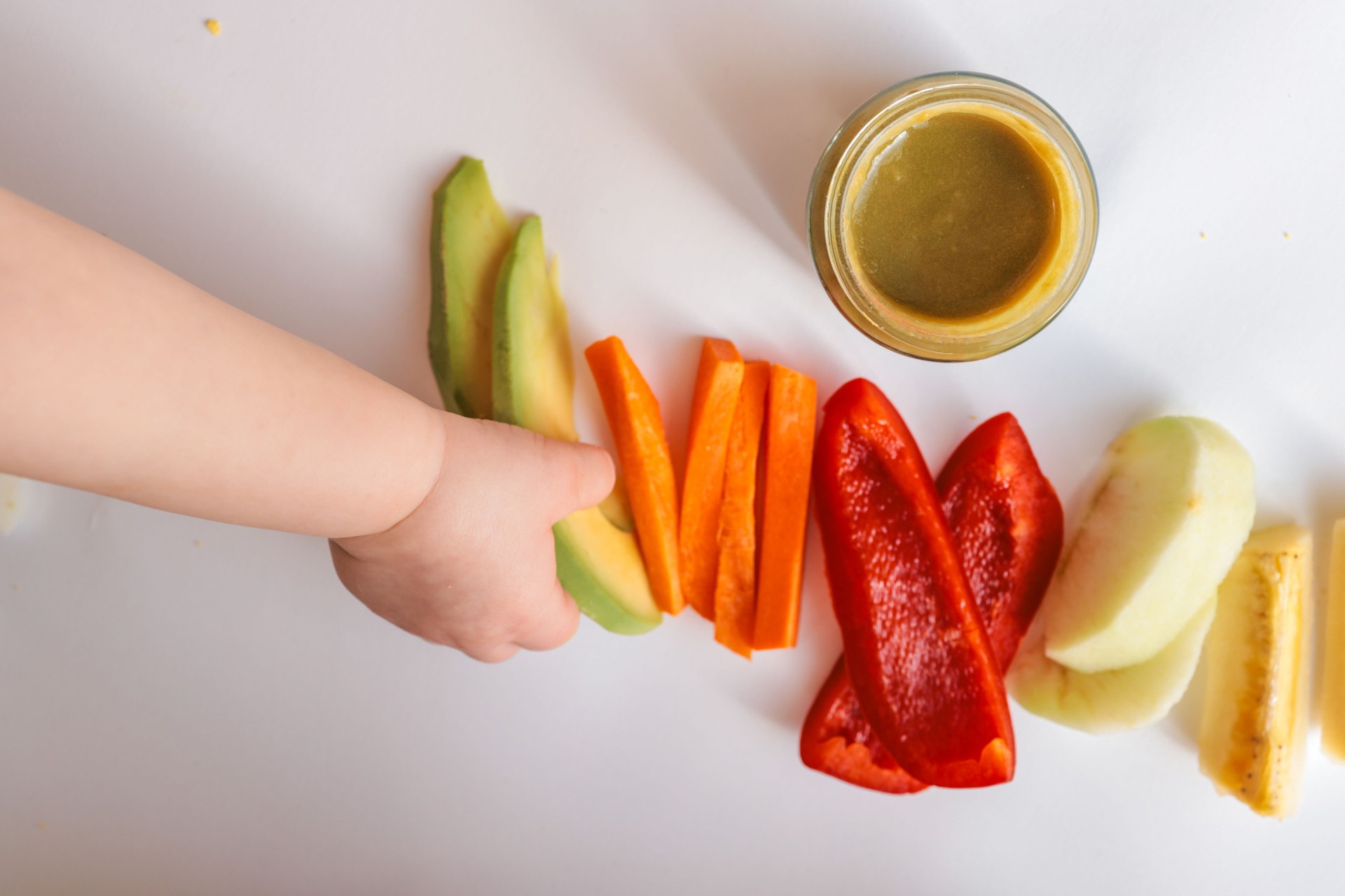 A child's hands reaching for freshly chopped vegetables on a white surface. Includes diced avocado, carrot, apple slices, bell pepper, and tomato pieces. Minimalist food composition.